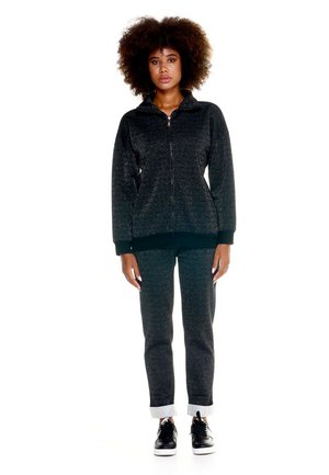 Une femme porte un sweat à capuche gris foncé zippé, un pantalon assorti à bas resserrés et des baskets noires, sur fond blanc.