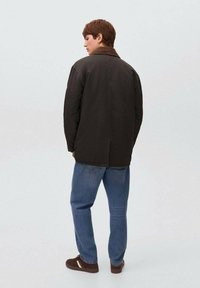 Giacca marrone scuro con colletto, caratterizzata da una forma squadrata e spalle scese, abbinata a jeans blu e scarpe marroni. Sfondo semplice.