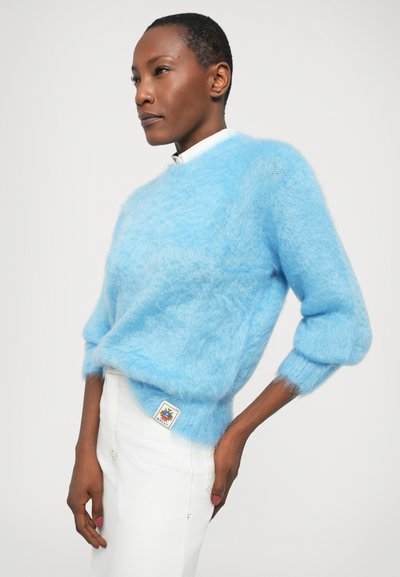 Pull en laine bleu clair, doux et duveteux, avec un col rond, des épaules tombantes et des manches trois-quarts, associé à un pantalon blanc orné d'une étiquette décorative.