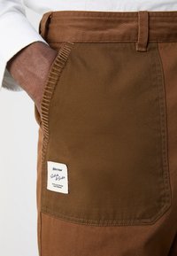 Brown cotton pants with a side pocket, featuring a stitched accent and a branded label with blue text. Smooth, sturdy fabric texture.
