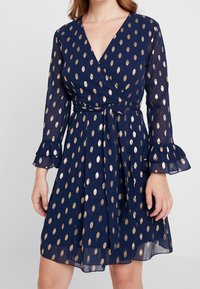 Marinblå omslagklänning med guldfolie ovalmönster, långa ärmar och volangärmar. Lätt, flödande tyg med detalj av knytning i midjan.