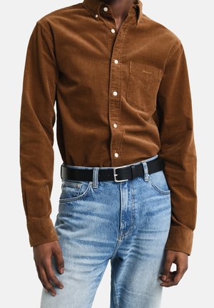 Brązowa koszula z korduroyu zapinana na guziki, z kieszenią na piersi, w zestawie z jasnoniebieskimi, wyblakłymi dżinsami i czarnym paskiem. Teksturowany materiał; dopasowany krój.