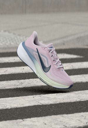 Zapatilla para correr Nike de color púrpura claro con el símbolo azul, en el aire sobre un paso de peatones en blanco y negro sobre el pavimento.