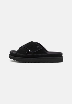 Sandalia tipo slide de ante negro con tiras entrecruzadas, suela acolchada y logo de la marca "UGG" en la tira lateral.