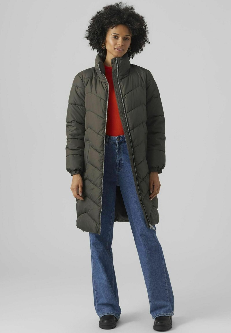 Lange, olijfgroene pufferjas met een hoge kraag; voorzien van een rits aan de voorkant en een ruitvormig doorstikpatroon. Gecombineerd met blauwe wijde jeans.