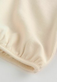 Jasnobeżowa tkanina o gładkiej fakturze, z marszczonym brzegiem i prążkowanym wykończeniem. Materiał wydaje się miękki i przewiewny.