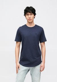 Marinblå bomullst-shirt med rund halsringning, korta ärmar och en liten svart logotyp på bröstet. Kombinerad med ljusa denimjeans.