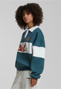 Pulover cu dungi teal și alb, cu guler, având un grafic brodat al unei creaturi cu aripi. Material moale cu manșete cu țesătură rib.