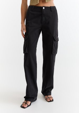 Kobieta w czarnych spodniach typu cargo z klapkowymi kieszeniami i czarnych sandałach na paski, stojąca na białym tle.