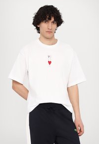 Tricou alb din bumbac, croi lejer, cu un grafic colorat în formă de inimă și cifre pe piept. Asortat cu pantaloni închiși la culoare.
