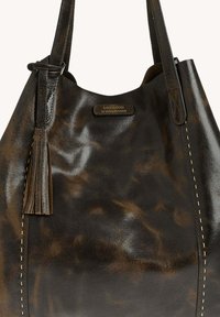 Sac à main en cuir marron foncé avec des bords cousus, un pompon sur une sangle et une petite plaque logo en lettres dorées à l'avant.