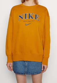Orange bomullströja med en stor blå "NIKE"-logotyp och texten "Athletic dept. 1972" över bröstet. Ribbad mudd och nederkant.