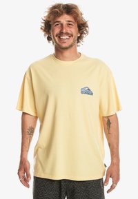 Camiseta de algodón amarilla con mangas cortas, cuello redondo, y un gráfico de logotipo azul en el lado izquierdo del pecho. Se lleva con pantalones cortos con estampados.