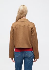 Brun kort jakke i imiteret ruskind med hakket krave og lange ærmer, båret over en rød skjorte, kombineret med blå cowboybukser.