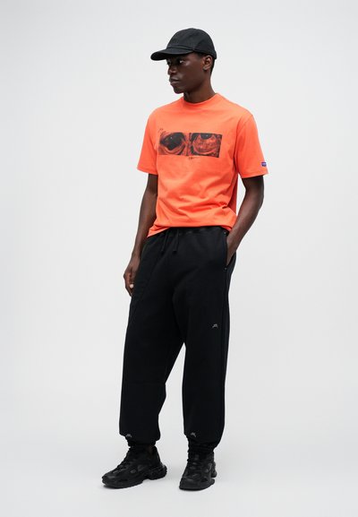 Oranžové bavlnené tričko s grafickým potlačou, spárované s voľnými čiernymi teplákmi. Čierne tenisky a čiapka dokončujú outfit.
