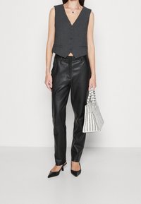 Graue ärmellose Weste mit V-Ausschnitt und vorderen Knöpfen, kombiniert mit schwarzen Lederhosen und einer weißen gestreiften Handtasche. Schwarze spitze High Heels vervollständigen den Look.