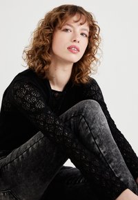 Haut en dentelle noire à manches longues avec motif floral, associé à un jean slim délavé gris. Modèle assis, montrant les détails et textures du tissu.