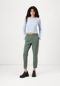 Světle modrý žebrovaný top s dlouhým rukávem a vlnitým lemem, kombinovaný s elegantními zelenými kalhotami; model má na sobě černé dřeváky a stojí na bílém pozadí.