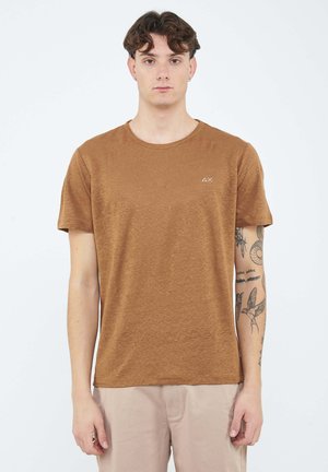 Giovane uomo con capelli corti e ricci che indossa una maglietta marrone a maniche corte e pantaloni beige, con tatuaggi visibili sul braccio destro.