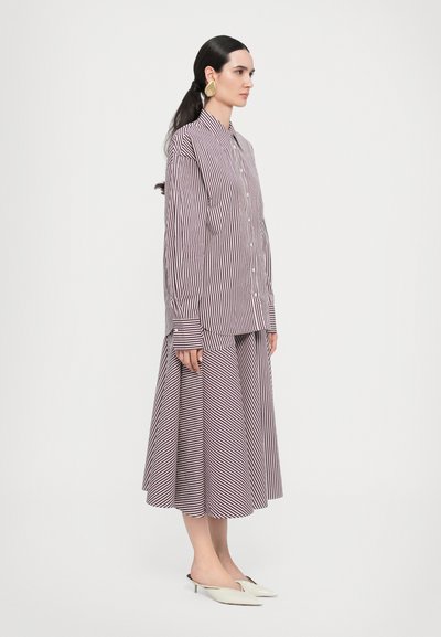 Abito a righe in un profondo bordeaux e bianco, con parte superiore a maniche lunghe e bottoni e una gonna fluida a forma di A. Scarpe slip-on chiare.