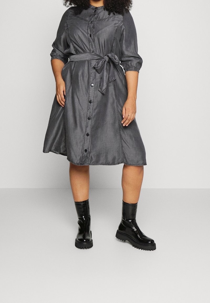 Grå klänning med knapplist fram, knytmidja och 3/4-långa ärmar. Slät textur, utsvängd form, ihop med glänsande svarta ankelstövlar.