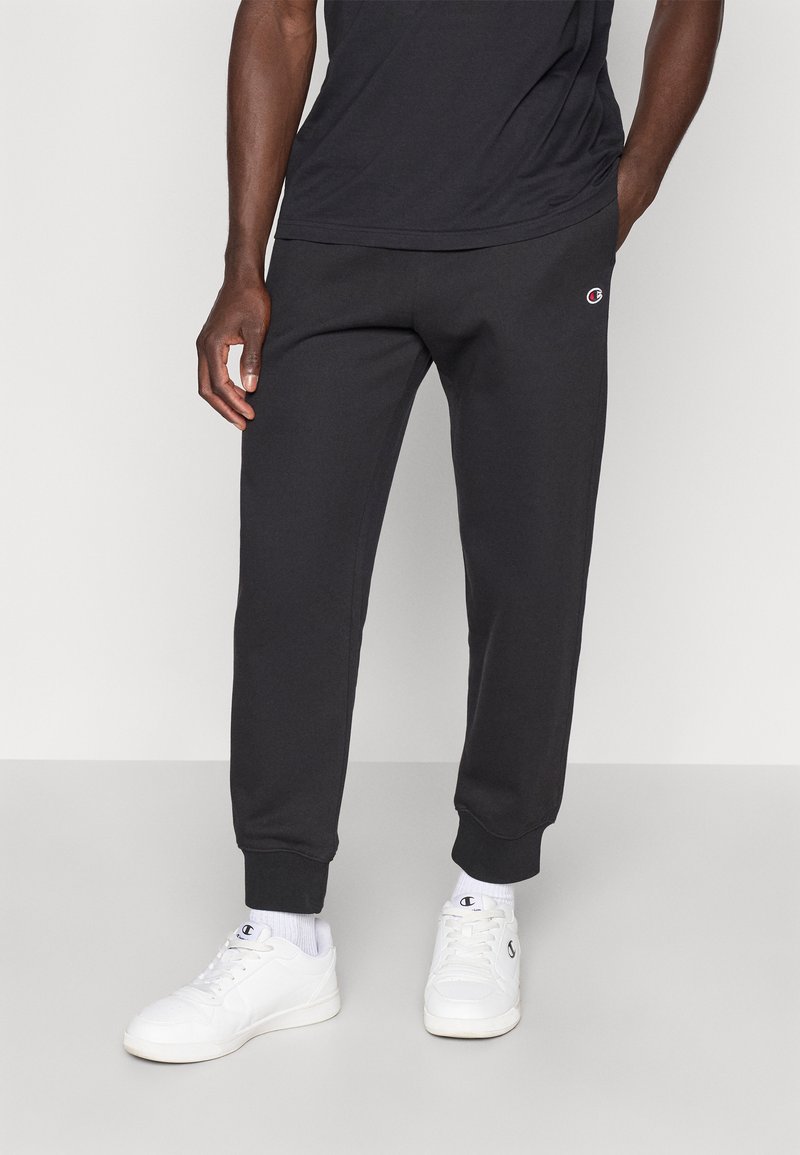 Μαύρο παντελόνι φόρμας με ριμπ στη μέση και μανσέτες στα πόδια, φτιαγμένο από απαλό ύφασμα. Διαθέτει μικρό λογότυπο στο αριστερό μηρό. Φοριέται με λευκά αθλητικά παπούτσια.