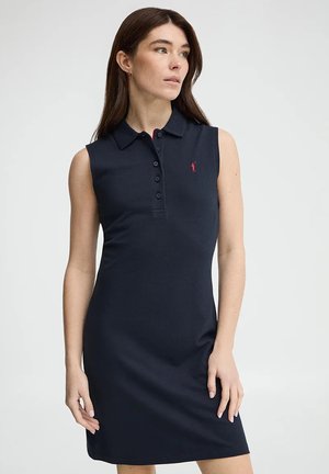 Donna con capelli castani lunghi che indossa un abito polo senza maniche blu navy con bottoni e un piccolo logo rosso ricamato, che guarda di lato.