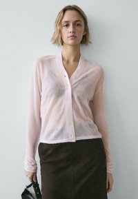 Cardigan rose clair transparent avec un col en V et une fermeture boutonnée, associé à une jupe marron foncé. Le tissu semble doux et texturé.