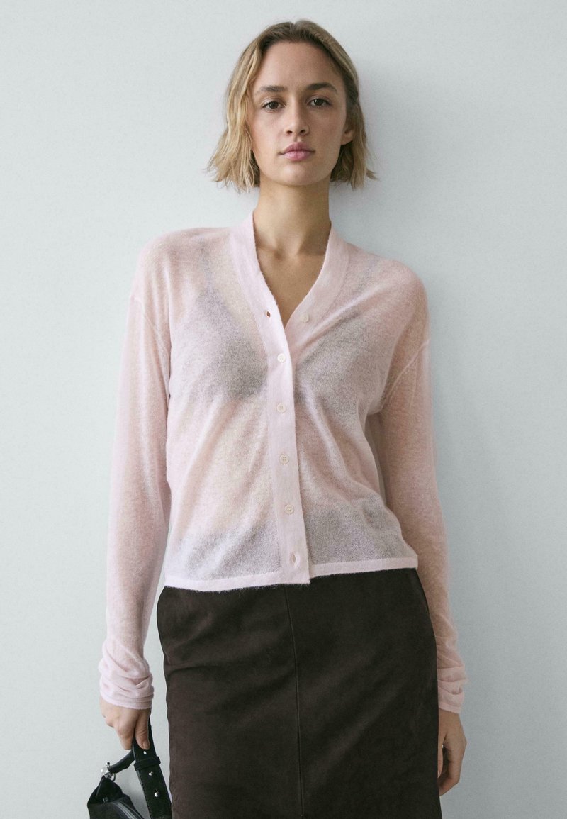 Cardigan rose clair transparent avec un col en V et une fermeture boutonnée, associé à une jupe marron foncé. Le tissu semble doux et texturé.