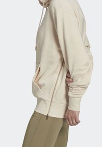 Beige hoodie med dragkedjeförsedd ficka, dragsko i huvan och mjukt tyg. Kombinerad med olivgröna texturerade leggings med en logotyp.