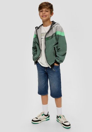 Groene en grijze windbreaker met zwarte accenten, gecombineerd met blauwe denim shorts. Een witte T-shirt en zwarte schoenen met teal accenten maken de look compleet.