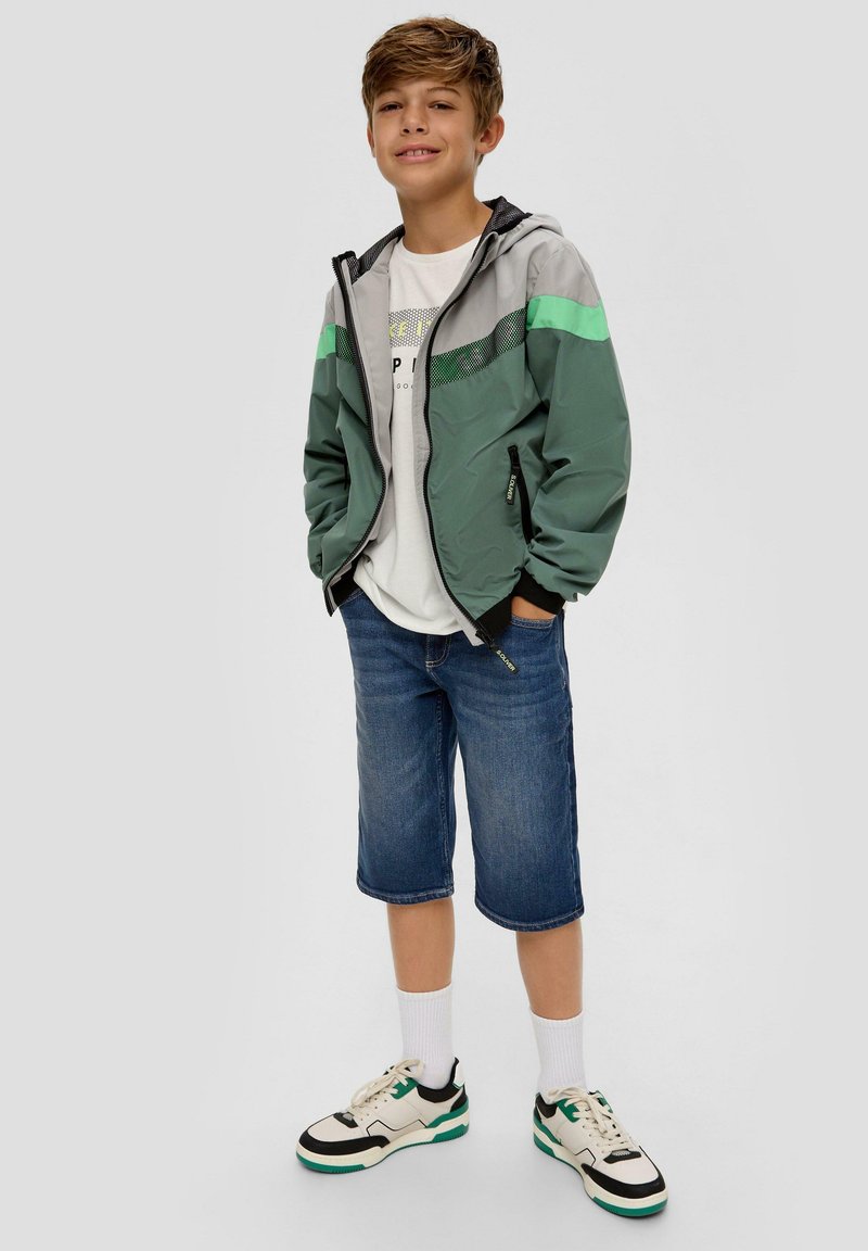 Coupe-vent vert et gris avec des bordures noires, associé à un short en denim bleu. Un t-shirt blanc et des chaussures noires avec des accents teal complètent le look.