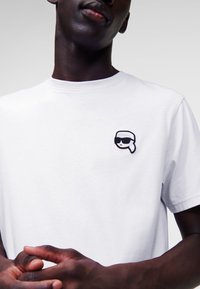 Witte katoenen t-shirt met een kleine zwart-wit geborduurde afbeelding van een persoon met een zonnebril op de linkerborst.