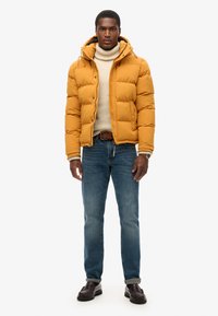 Giacca a vento gialla con un design aderente, cappuccio e cuciture in vista. Indossata sopra un maglione chiaro, abbinata a jeans blu e scarpe scure.