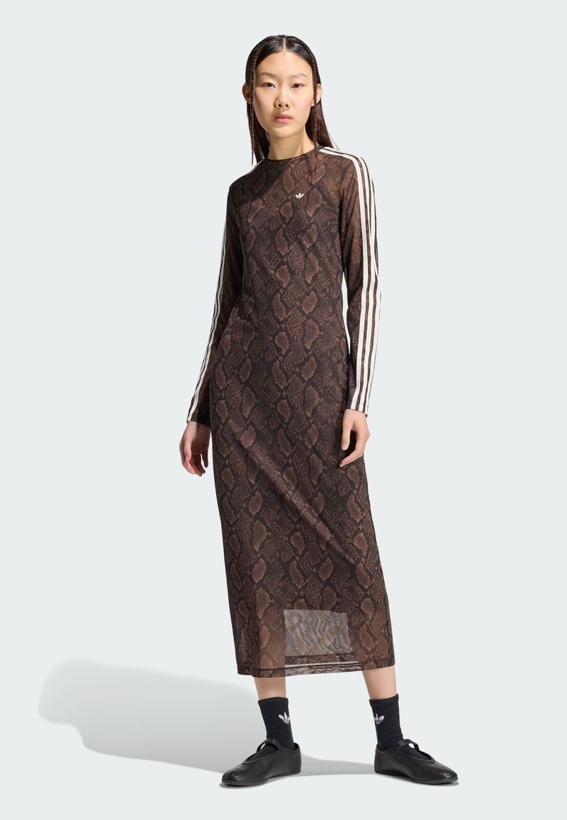 Femme portant une longue robe marron à motif peau de serpent avec des rayures blanches sur les manches, des chaussettes noires et des chaussures noires, debout sur un fond uni.