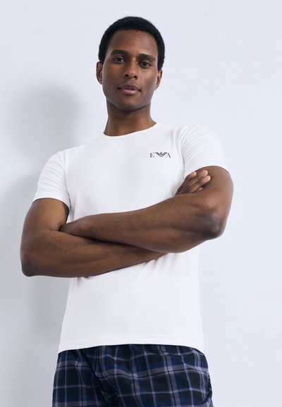 Bílé přiléhavé tričko z hladké tkaniny s malým černým logem na levé straně hrudi. Model pózuje se založenýma rukama a má na sobě kostkované pyžamové šortky.