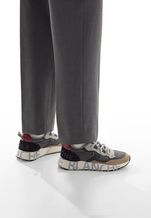 Persona con pantaloni grigi e scarpe da ginnastica nere, grigie e beige con grandi testi "BLA" e "NCHE" sulle suole, in piedi su un pavimento bianco.