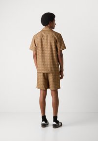 Men's brown patterned short-sleeve shirt and matching shorts, featuring geometric designs, standard collar, and relaxed fit. Black socks and shoes.