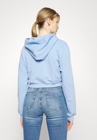 Sweatshirt cropped azul claro com capuz ajustável, apresentando punhos e barra canelados. Combinado com jeans de lavagem média.