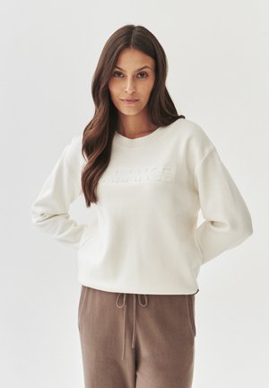 Cremfarbener Sweatshirt mit strukturiertem "BALANCE"-Logo, Rundhalsausschnitt und legerer Passform. Kombiniert mit braunen, mit Kordelzug versehenen Hosen. Weicher Stoff.
