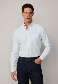 Lichtblauw gestreept overhemd met knopen en een zak, gedragen over een roze onderhemd, gecombineerd met donkerblauwe spijkerbroek.