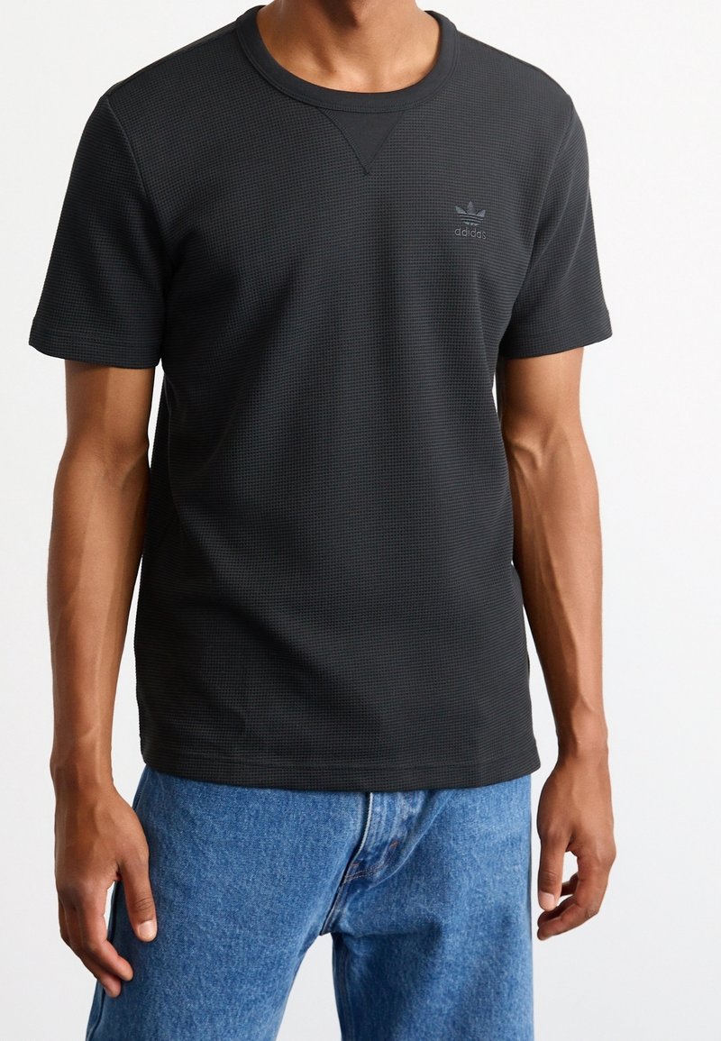 Svart kortärmad t-shirt med en texturerad mönster, rund halsringning, och adidas-logotyp i en mörkare nyans på bröstet. Bärs tillsammans med ljusblå jeans.