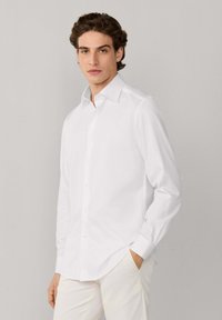 Jeune homme aux cheveux bruns ondulés portant une chemise blanche impeccable à boutons et un pantalon clair, debout devant un fond clair uni.