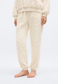 Κοντινό πλάνο ενός ατόμου που φοράει υφασμάτινο παντελόνι φλις σε κρεμ απόχρωση με ριμπ μανσέτες, στέκεται ξυπόλυτο σε λευκό φόντο.