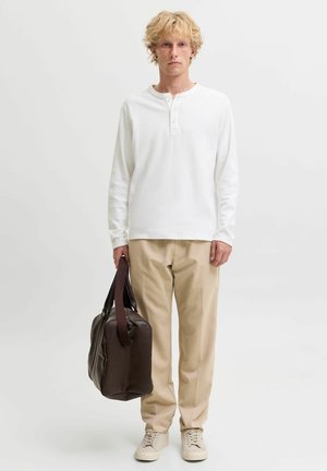 Giovane uomo con capelli biondi e ricci che indossa una camicia bianca a maniche lunghe e pantaloni beige, tenendo una borsa in pelle marrone scuro nella mano destra.