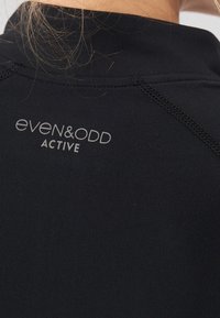 Schwarzes Aktivwear-Oberteil mit hohem Kragen, glatter Textur und flachen Nähten. Der Markenname "even&odd ACTIVE" ist in Grau aufgedruckt.