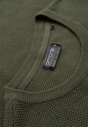 Olivgrüner Pullover mit strukturiertem Strickmuster, geripptem Kragen und sichtbarem Etikett, das den Markennamen und Materialdetails hervorhebt.