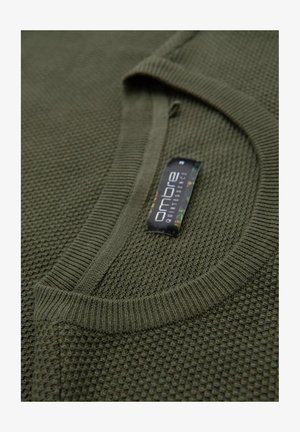 Olivgrüner Pullover mit strukturiertem Strickmuster, geripptem Kragen und sichtbarem Etikett, das den Markennamen und Materialdetails hervorhebt.