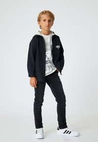 Zwarte spijkerjacket over een wit graphic T-shirt, gecombineerd met zwarte spijkerbroeken en witte sneakers met zwarte strepen. Eenvoudig, modern ontwerp.