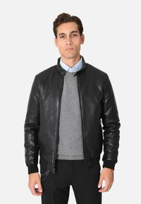 Giubbotto bomber in pelle nera con zip frontale, polsini a coste e colletto rialzato. Indossato sopra un maglione grigio e abbinato a pantaloni neri.